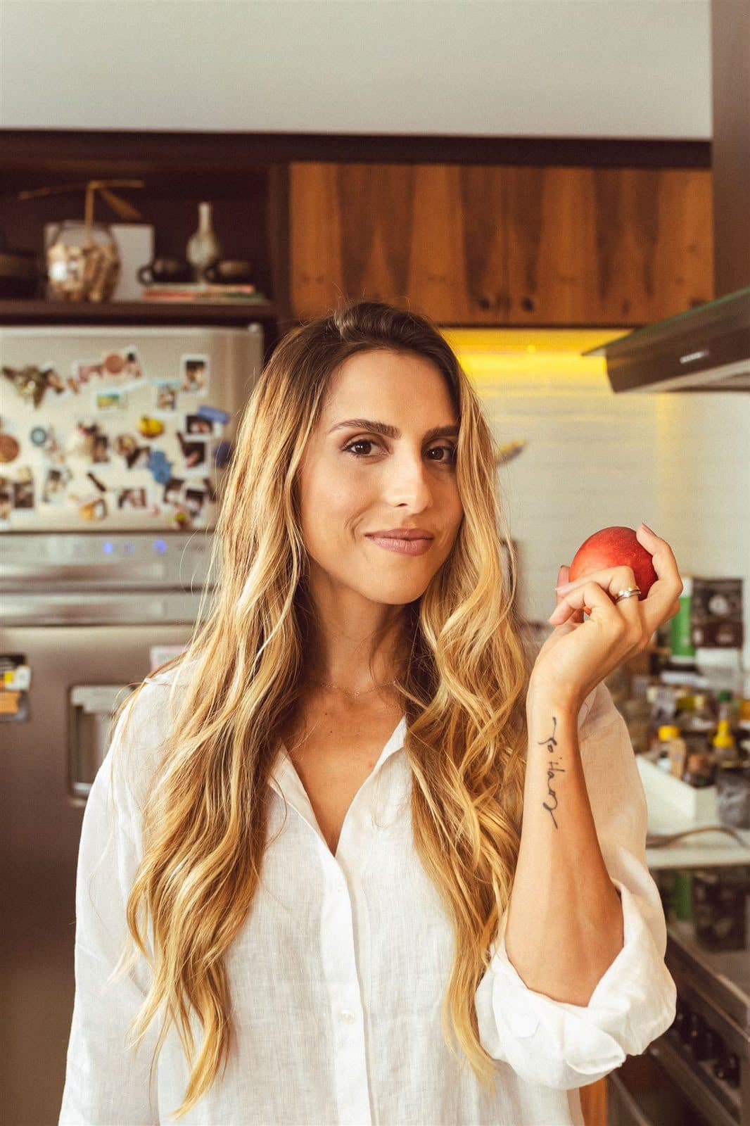 Xanda Fogaça na cozinha, de camisa branca, segurando uma fruta